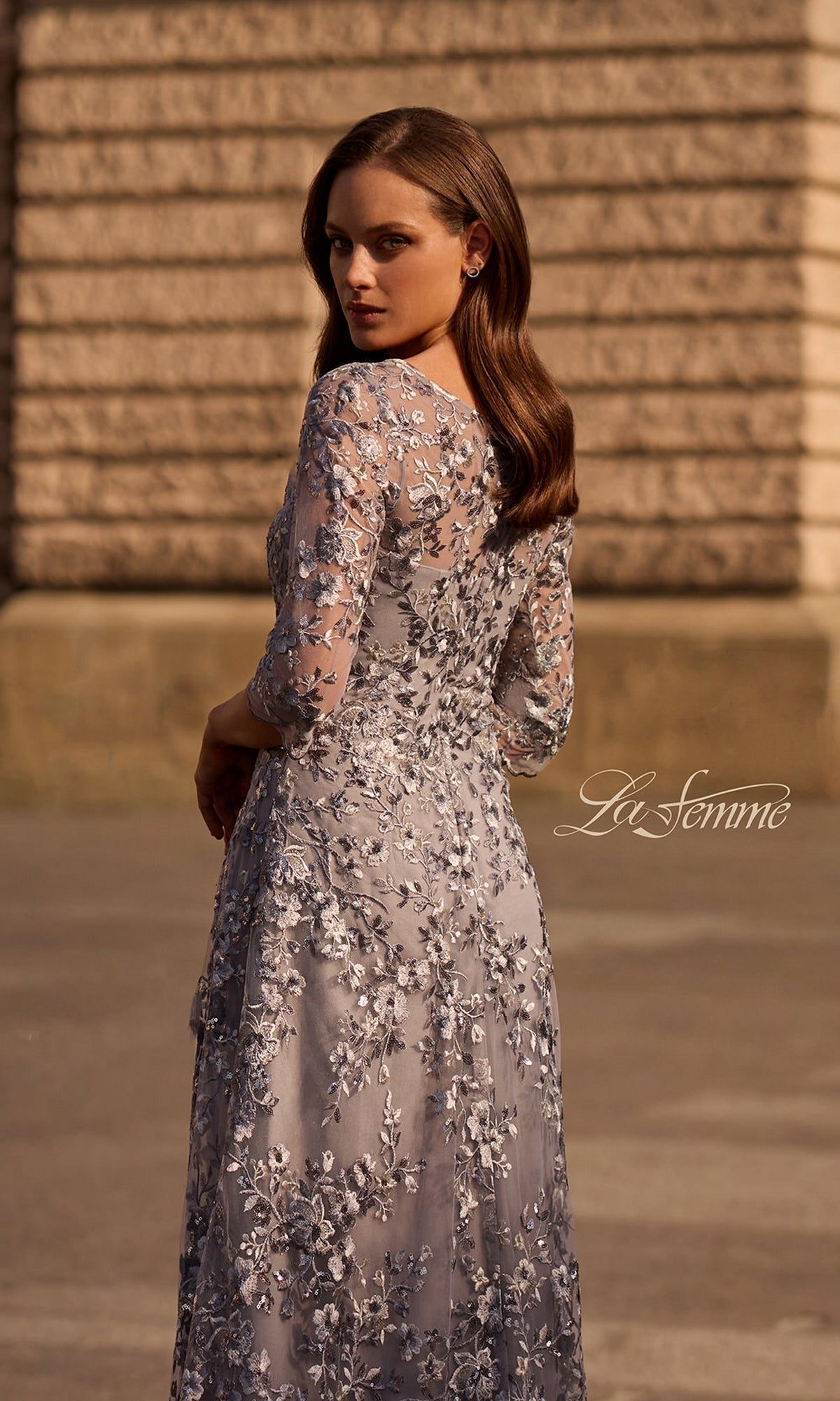 A woman wearing the Long Formal Dress: La Femme 29903, a silver modest gown with metallic floral lace embroidery, stands outdoors looking over her shoulder.